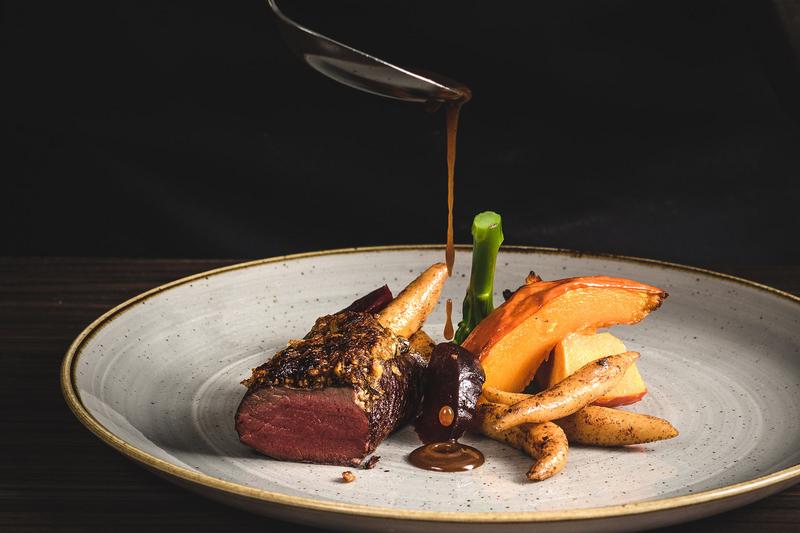 Ein Koch gießt Sauce mit einem Löffel über ein Gericht aus Fleisch und Gemüse